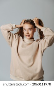 Confident Pretty Teenage Girl With Brackets Holding Her Hair In Pony Tails. Against Grey Background. In A Warm Beige Longsleeve.