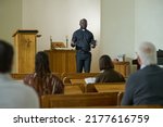 Confident priest of evangelical church with Holy Bible in hand saying sermon while standing in front of intercultural parishioners