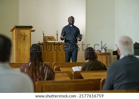 Confident priest of evangelical church with Holy Bible in hand saying sermon while standing in front of intercultural parishioners