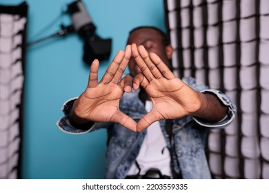 Confident producer with professional camera posing beside softboxes and spotlights. Smiling photography enthusiast making love symbol with hands while standing in production studio. - Shutterstock ID 2203559335