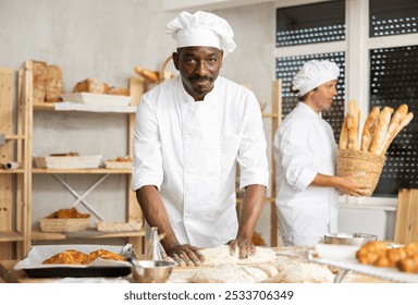 Confident professional African American male baker smiling and carefully shaping dough into baguette on floured table, preparing fresh bread in cozy artisan bakery - Powered by Shutterstock