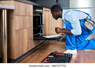 Confident professional afro american guy is looking inside of oven, going to repair, wearing blue workwear uniform, Side view portrait. Handyman of african appearance sit on floor in kitchen - Shutterstock ID 2067577751