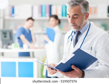 Confident Professional Doctor In The Office Checking Patient's Medical Records On A Clipboard, Medical Staff Working On The Background