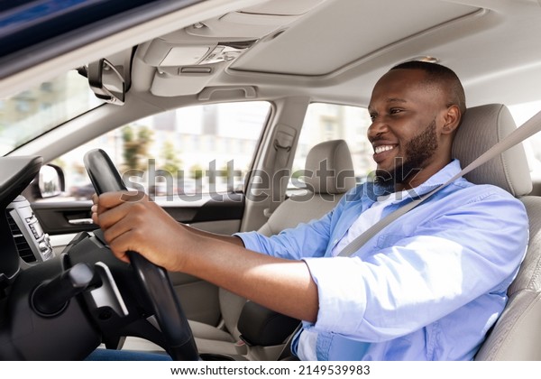 Confident Professional Driver. Side view profile\
portrait of smiling young African American man sitting in a car on\
driver\'s seat. Positive black guy riding in the city holding hands\
on steering wheel
