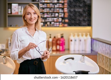confident professional hairdresser in beauty saloon, caucasian woman with blond hair wearing white shirt stand looking at camera, isolated in beauty saloon. Cosmetics, balms and shampoos in background