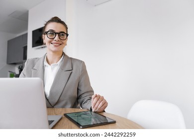 A confident professional woman in a chic jacket smiles while working on a digital tablet, seated at her desk in a minimalist office.

 - Powered by Shutterstock