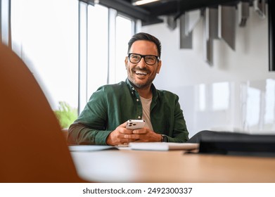 Confident project manager in glasses using smartphone and looking away while sitting at desk in office - Powered by Shutterstock