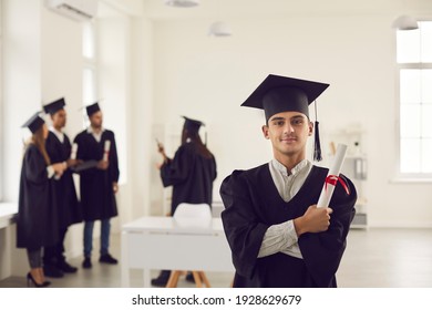 Confident And Proud Student In The Form Of A University Graduate And With A Diploma In Hand Stands In A Bright Classroom. Portrait Of A Guy Posing With Crossed Arms Against The Backdrop Of Classmates.