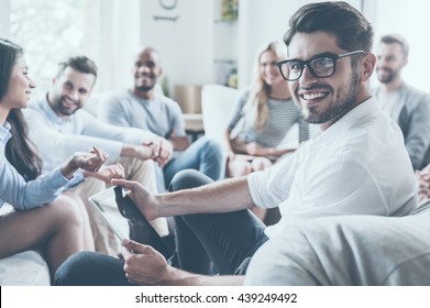 Confident psychologist. Group of young cheerful people sitting in circle and discussing something while young man holding digital tablet and looking over shoulder with smile - Powered by Shutterstock