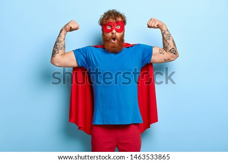 Confident red head male superhero wears mask and red cape, raises arms, shows muscles, represents power and courage, ready to defend people, has shocked expression, isolated on blue background