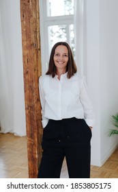 Confident relaxed professional woman standing with hands in pockets leaning on an interior wooden post smiling at the camera