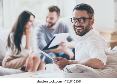 Confident Sales Manager. Cheerful Mature Man Holding Digital Tablet And Looking Over Shoulder While Another Man And Woman Sitting In The Background  