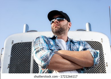 Confident semi truck driver wearing plaid shirt and black baseball cap  stands with arms crossed in front of big rig. Portrait of professional looking trucker. Blue collar jobs in transportation. 