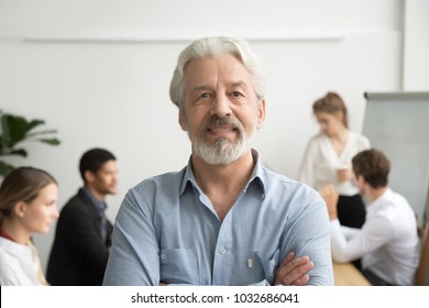 Confident Senior Businessman Leader Looking At Camera With Team At Background, Smiling Aged Company Boss Posing In Office With Arms Crossed, Older Mentor Or Executive Professional Head Shot Portrait