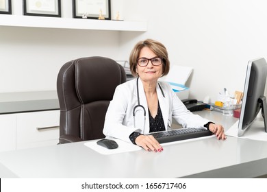 Confident Senior Female Doctor Sitting At Computer Desk In Office