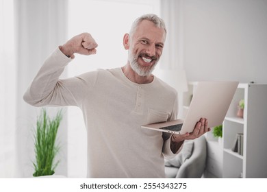 Confident senior man celebrating success while holding laptop in comfortable home environment - Powered by Shutterstock