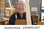 Confident senior man, a professional carpenter, smiling while securely holding a wood plank in carpentry workshop