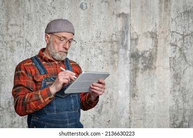 Confident Senior Man Wearing Glasses, A Flannel Shirt And Overalls Is Making Notes Or Sketches On A Digital Tablet