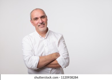 Confident senior man in white t-shirt crossing hands on chest and looking at camera while standing against gray background. Self confident hispanic senior.