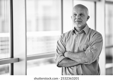 Confident senior manager stands in a modern office, arms crossed, looking out the window with a thoughtful gaze - Powered by Shutterstock