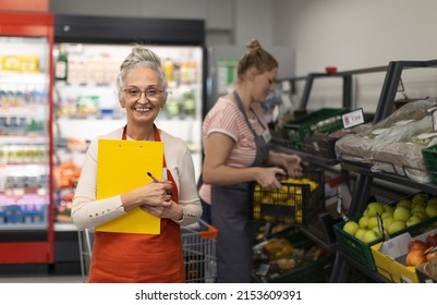 Confident senior shop assistant in supermarket in vegetable shell, in bakcground is her colleague filling stock. - Powered by Shutterstock