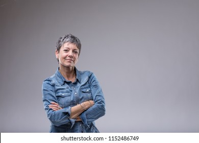 Confident senior woman with folded arms standing looking at camera with a thoughtful quiet smile over grey with copy space