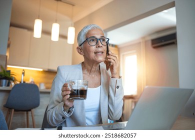 Confident senior woman multitasking at home office with a smartphone and coffee, showcasing modern technology and work-life balance in a cozy kitchen setting. - Powered by Shutterstock