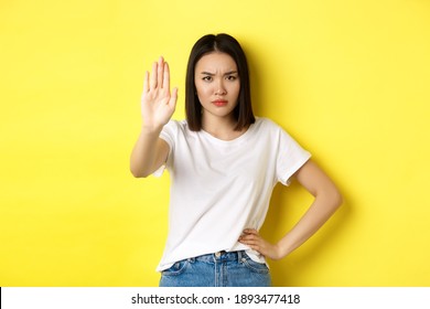 Confident And Serious Asian Woman Tell No, Showing Stop Gesture To Prohibit And Warn, Disagree With Someone, Standing Upset Against Yellow Background