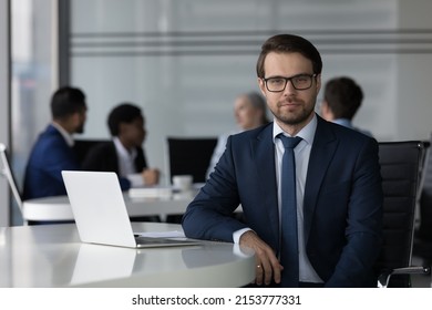 Confident Serious Business Leader Man In Glasses Corporate Portrait With Team Of Coworkers Meeting In Background. Businessman Working At Laptop In Co-working Space, Looking At Camera