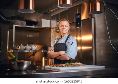 Confident and serious female chef standing with hands crossed in a dark kitchen next to cutting board with vegetables on it, wearing apron and denim shirt, looking in the camera, cooking show look