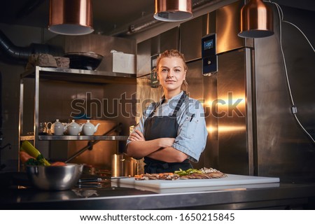 Confident and serious female chef standing with hands crossed in a dark kitchen next to cutting board with vegetables on it, wearing apron and denim shirt, looking in the camera, cooking show look