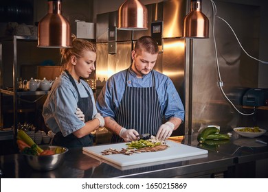 Confident And Serious Male And Female Chefs Standing In A Dark Kitchen Next To Cutting Board With Vegetables On It, Wearing Aprons And Denim Shirts, Posing For The Camera, Cooking Show Style, Teaching