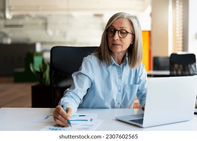 Confident serious mature businesswoman using laptop computer, taking notes, working  project in modern office. 60 years old female, financier working with documents at workplace. Successful business  - Shutterstock ID 2307602565