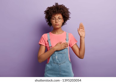 Confident serious woman with curly hair makes sincere promise or oath, keeps one hand on heart, solemnly swears, raises palm, demonstrates loyalty gesture being honest poses against purple background.