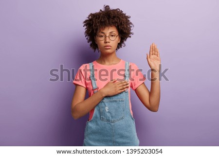 Confident serious woman with curly hair makes sincere promise or oath, keeps one hand on heart, solemnly swears, raises palm, demonstrates loyalty gesture being honest poses against purple background.