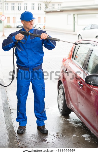 Confident serviceman with high pressure water jet\
washing car at service\
station