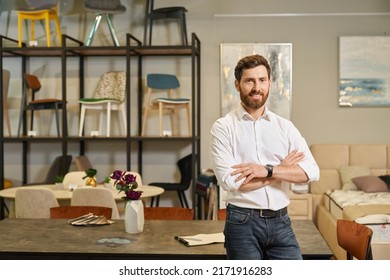 Confident Shop Owner In White Shirt Posing, With Crossed Arms In Furniture Store. Portrait Of Handsome Businessman Looking At Camera, Smiling, While Leaning On Table In Showroom. Concept Of Business.