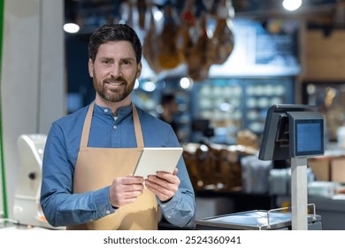 Confident shopkeeper wearing apron holding tablet in hands at store counter with digital scale. Professional retail worker surrounded by fresh produce, showcasing modern technology in business. - Powered by Shutterstock