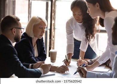 Confident Skilled Middle Aged Female Boss Making Remarks In Paper Marketing Research Report At Brainstorming Meeting. Focused Group Of Mixed Race Young Managers Listening To Leader Remarks At Office.