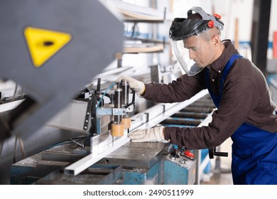 Confident skilled workman in blue overalls, protective face shield and gloves cutting PVC profile on industrial machine in window production and assembly workshop.. - Powered by Shutterstock