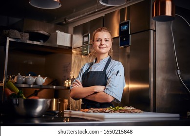 Gewissene und leicht lächelnde Küchenchefin, die in einer dunklen Küche neben dem Schneiden mit Gemüse darauf steht, mit Schürze und Denim-Shirt, posiert für die Kamera, Reality-Show-Look