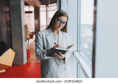 Confident slovenian business woman in elegant clothes checks notes in a notebook standing by the window in the office, a serious female manager reads information viewed on a notebook