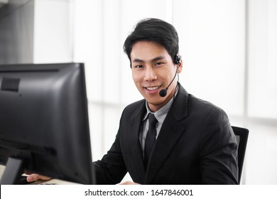 Confident Smiling Asian Business Man Call Center Staff At Office Desk Talking On Phone In A Call Centre, Portrait Of Male Operator Agent With Headsets, Customer Service Or Technical Support Concept