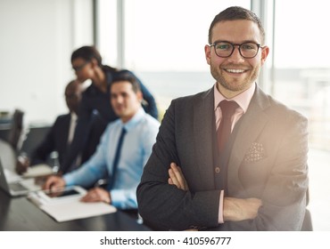 Confident smiling business executive with folded arms near conference table with three co-workers discussing something in large bright office room