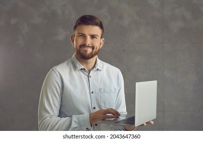 Confident Smiling Business Expert Working With Joy. Millennial Caucasian Handsome Man In Shirt Holding Laptop Looking At Camera With Smile Standing Against Studio Copy Space. People Emotion Concept