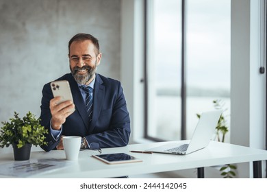 Confident smiling businessman using smartphone in office. Smart adult man, working hard in the office, checking his notes.Handsome businessman sitting in modern office.  - Powered by Shutterstock