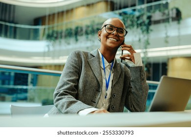 A confident and smiling businesswoman in a modern office environment, using her phone while sitting at a desk with her laptop. She wears professional attire, exuding positivity and success. - Powered by Shutterstock