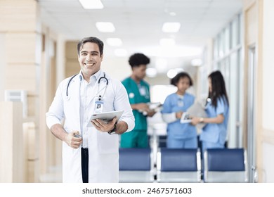 Confident smiling doctor posing and the hospital with arms crossed and medical team working on the background - Powered by Shutterstock