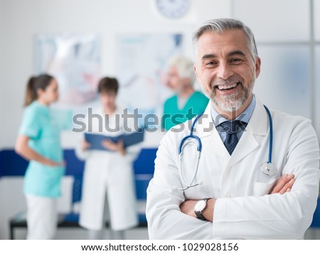 Confident smiling doctor posing and the hospital with arms crossed and medical team working on the background