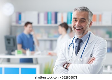 Confident smiling doctor posing and looking at camera with arms crossed, medical staff working on the background - Shutterstock ID 382908871
